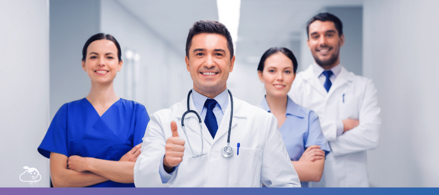 Grupo de quatro profissionais de saúde sorrindo em um corredor de hospital, com o médico central fazendo um sinal de positivo, simbolizando sucesso e confiança nas frases para medicina sobre excelência e trabalho em equipe.