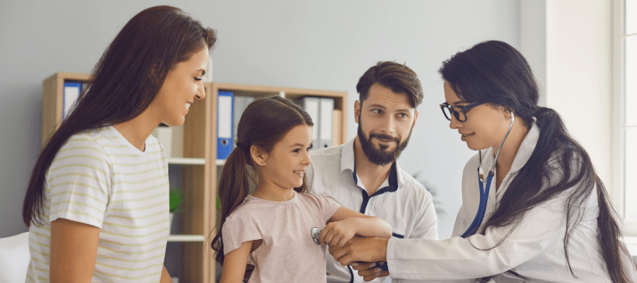 Médica de óculos e jaleco examinando uma menina sorridente com um estetoscópio, enquanto os pais observam com carinho no consultório, ilustrando o cuidado e a humanização nas frases para medicina pediátrica.