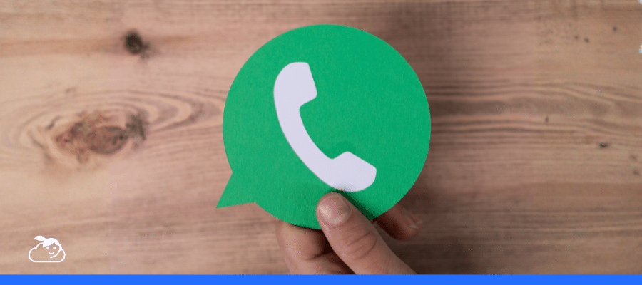 Mão segurando um recorte de papel verde que representa o ícone do WhatsApp (balão de fala e telefone) sobre uma superfície de madeira.