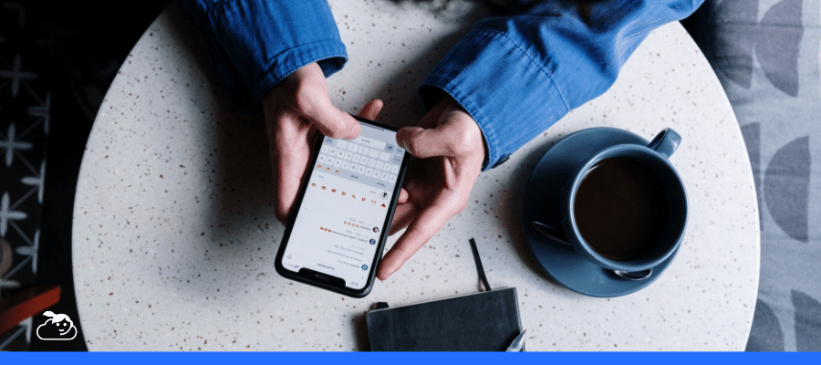 Close-up de uma pessoa digitando em um smartphone sobre uma mesa, com o teclado de emojis aberto e uma xícara de café ao lado, simbolizando a comunicação digital e interativa com um chatbot na saúde em um ambiente casual.