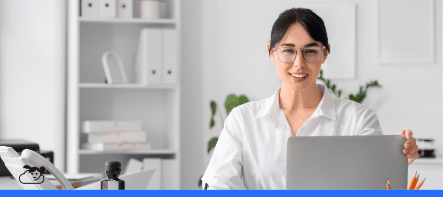 Secretária sorridente, de óculos e camisa branca, sentada à mesa com um laptop, telefone e carimbo, simbolizando a organização e a gestão de dados com um CRM para clínicas.