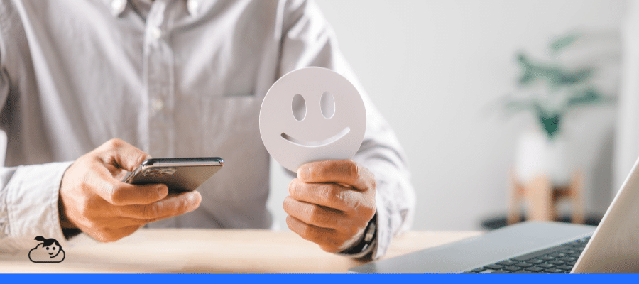 Homem sentado à mesa segura um smartphone em uma mão e um cartão com o desenho de um rosto sorridente na outra, simbolizando a avaliação de métricas de experiência do paciente em canais digitais. Ao fundo, um ambiente de escritório iluminado com um notebook e uma planta.