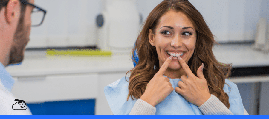 Paciente jovem sentada na cadeira odontológica demonstra satisfação com o resultado do tratamento. Ela sorri e aponta para o sorriso, evidenciando o sucesso da experiência do paciente na odontologia.